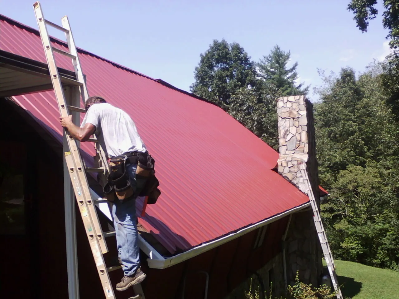 Roofer on a ladder beside a red metal roof and stone chimney — Soffit & Fascia Repair by Roofing Brothers in White Earth